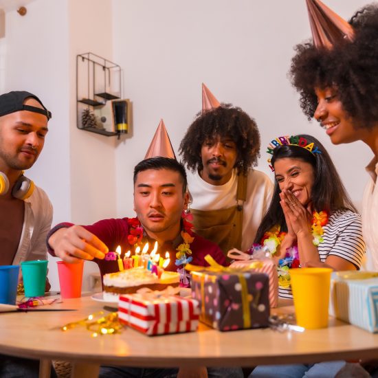 Multi-ethnic group of friends at a birthday party on the sofa at home with a cake and gifts, finishing lighting the candles