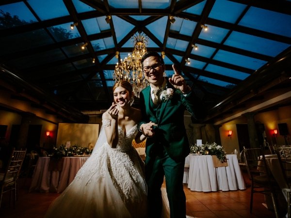A bride and groom celebrating under a grand chandelier in a glass-roofed wedding venue in Quezon City.